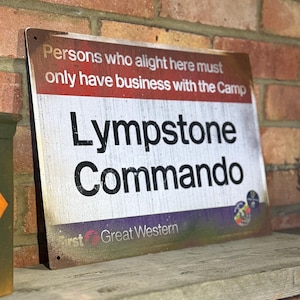 May include: A weathered metal sign with the words "Lympstone Commando" in large black letters. The sign also states "Persons who alight here must only have business with the Camp." It is mounted on a wooden shelf against a brick wall.