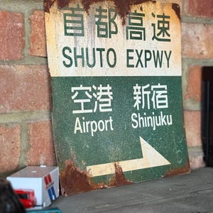 Könnte beinhalten: Ein verwittertes Metallschild mit japanischem Text und den englischen Wörtern "SHUTO EXPWY", "Airport" und "Shinjuku". Das Schild hat einen grünen und weißen Hintergrund mit einem Richtungspfeil. Das Schild ist stark verrostet und wirkt dadurch vintage.