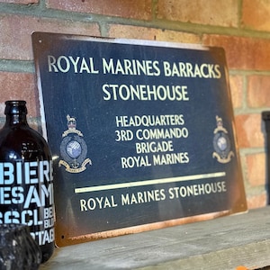 May include: A weathered metal sign with the text "ROYAL MARINES BARRACKS STONEHOUSE" in white. It also displays "HEADQUARTERS 3RD COMMANDO BRIGADE ROYAL MARINES." A dark bottle is visible to the left of the sign.
