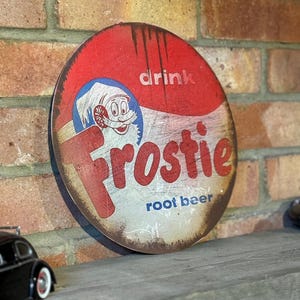 May include: Round, vintage-style metal sign with a red and white color scheme. The sign features the word "Frostie" in red, a cartoon character, and the words "root beer" and "drink". The sign has a distressed, aged appearance.