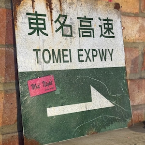 Puede incluir: Un letrero metálico desgastado con caracteres japoneses y el texto en inglés "TOMEI EXPWY" y una flecha blanca. El letrero tiene un fondo verde y blanco con una pegatina "Mid Night Racing Spirit". El letrero tiene una apariencia vintage.