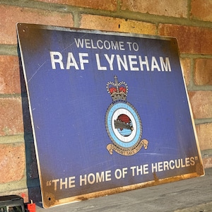 May include: A rectangular blue metal sign with the text "WELCOME TO RAF LYNEHAM" and "THE HOME OF THE HERCULES". The sign has a royal emblem and is displayed against a brick wall. A small green toy jeep is visible in the foreground.