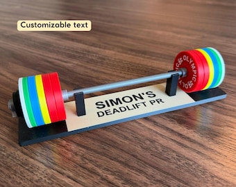 May include: A miniature barbell display stand featuring a grey bar with stacks of red, blue, yellow, and white weight plates on each end. The base is black with a beige plaque reading SIMON'S DEADLIFT PR. The text is customizable for this item.