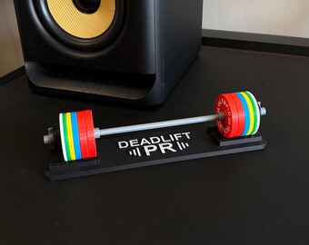 May include: A miniature barbell with colorful weight plates, including red, blue, yellow, and green. The barbell rests on a black base with the text "DEADLIFT PR" in white. The weights are labeled "OLYMPIC RX".