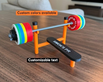 May include: A miniature orange weight bench with a black seat featuring the white text SIMON'S PR. A grey barbell rests on the rack, loaded with colorful red, yellow, blue, and white Olympic-style weight plates. The text Custom colors available and Customizable text appear in the image.