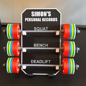 May include: A black and white sign displays "SIMON'S PERSONAL RECORDS" above three barbell designs. Each barbell has colourful weight plates in red, blue, green, and yellow. The sign lists "SQUAT", "BENCH", and "DEADLIFT".