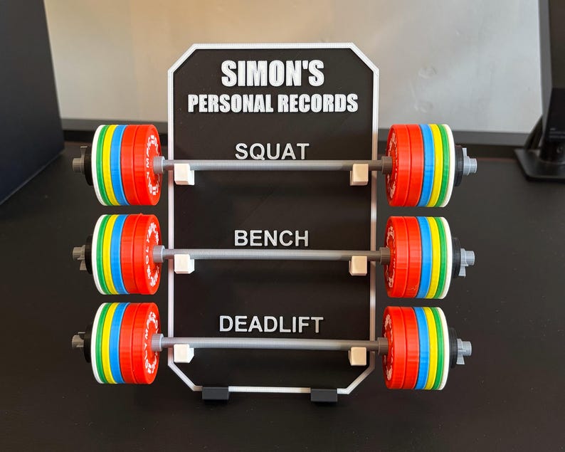 May include: A black and white sign displays "SIMON'S PERSONAL RECORDS" above three barbell designs. Each barbell has colourful weight plates in red, blue, green, and yellow. The sign lists "SQUAT", "BENCH", and "DEADLIFT".