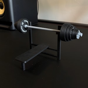 K&ouml;nnte beinhalten: Eine Miniatur-Hantelbank in Schwarz mit einer Langhantel und Gewichtsscheiben. Die Langhantel ist silberfarben, die Gewichte sind schwarz mit wei&szlig;er Schrift. Die Bank ist aus schwarzem Material. Die Gewichte sind mit "55 LBS" beschriftet.