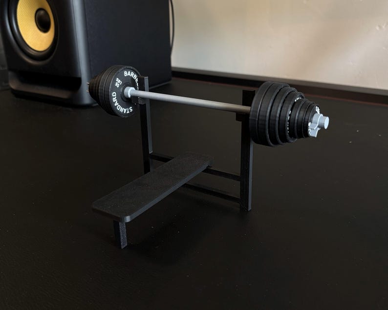 K&ouml;nnte beinhalten: Eine Miniatur-Hantelbank in Schwarz mit einer Langhantel und Gewichtsscheiben. Die Langhantel ist silberfarben, die Gewichte sind schwarz mit wei&szlig;er Schrift. Die Bank ist aus schwarzem Material. Die Gewichte sind mit "55 LBS" beschriftet.