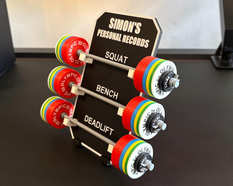 May include: A black sign displays "SIMON'S PERSONAL RECORDS" with three barbell sets labeled "SQUAT", "BENCH", and "DEADLIFT". Each barbell has red, yellow, blue, and green plates. The weights are marked in kilograms.