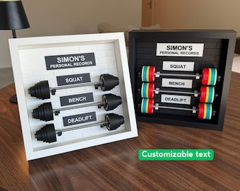 May include: Two square shadow box frames, one white and one black, each featuring a brick-patterned backboard. Inside are miniature barbells with weight plates and labels reading SIMON'S PERSONAL RECORDS, SQUAT, BENCH, and DEADLIFT.