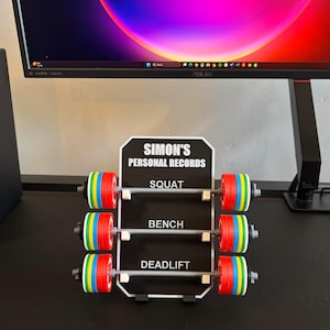 May include: A black and white sign reading "SIMON'S PERSONAL RECORDS" with three miniature barbell displays for Squat, Bench, and Deadlift. Each barbell has colourful weight plates.