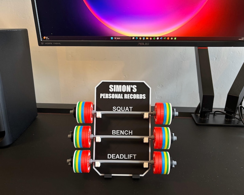May include: A black and white sign reading "SIMON'S PERSONAL RECORDS" with three miniature barbell displays for Squat, Bench, and Deadlift. Each barbell has colourful weight plates.