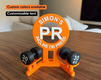 May include: An orange 3D-printed display stand featuring the white text SIMON'S PR INCLINE DB PRESS. The stand holds two miniature black dumbbells, each clearly marked with the weight 30 KG on the circular ends of the weights.