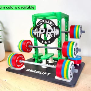 May include: A green plastic miniature squat rack display stand featuring a black circular sign with the text YOUR NAME and PR. The rack includes grey barbells with red, yellow, and blue weight plates labeled OLYMPIC. The base is black with the text DEADLIFT.