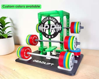 May include: A green plastic miniature squat rack display stand featuring a black circular sign with the text YOUR NAME and PR. The rack includes grey barbells with red, yellow, and blue weight plates labeled OLYMPIC. The base is black with the text DEADLIFT.