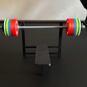 K&ouml;nnte beinhalten: Eine Miniatur-Hantelbank mit einer Langhantel und bunten Gewichtsscheiben. Die Langhantel ist silberfarben und die Gewichte sind rot, gelb, blau, gr&uuml;n und wei&szlig;. Die Bank und der Rahmen sind schwarz.