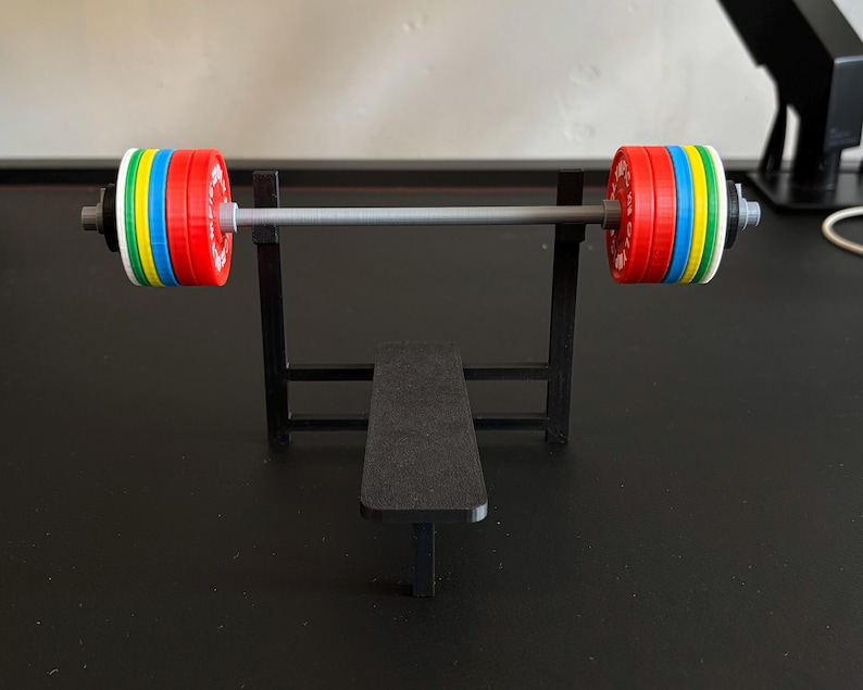 K&ouml;nnte beinhalten: Eine Miniatur-Hantelbank mit einer Langhantel und bunten Gewichtsscheiben. Die Langhantel ist silberfarben und die Gewichte sind rot, gelb, blau, gr&uuml;n und wei&szlig;. Die Bank und der Rahmen sind schwarz.