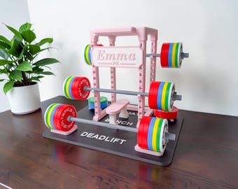 May include: A miniature pink weightlifting rack and barbell set on a black base with the text Emma, Bench, Squat, and Deadlift. The set includes grey barbells and colorful red, blue, green, and yellow weight plates labeled Olympic.