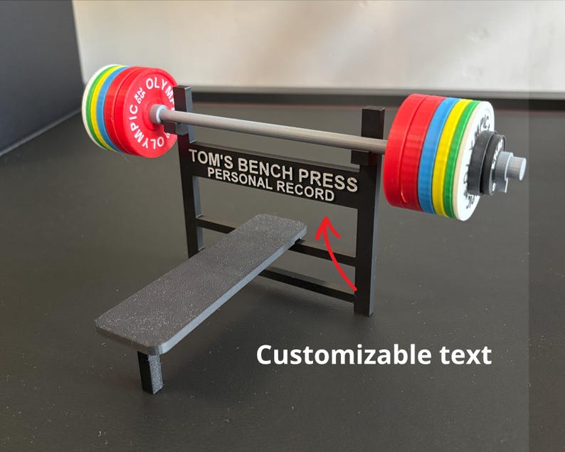 K&ouml;nnte beinhalten: Eine Miniatur-Gewichtheberbank mit Langhantel und Gewichtsscheiben. Die Langhantel hat rote, gelbe, blaue und gr&uuml;ne Scheiben. Auf der Bank steht der Text "TOM'S BENCH PRESS PERSONAL RECORD". Bank und Gewichte sind schwarz.