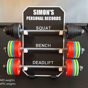 May include: A black and white sign displays "SIMON'S PERSONAL RECORDS" with three barbell sets for Squat, Bench, and Deadlift. The barbells have black or colourful weight plates. Text at the bottom reads "Choose STANDARD weights or OLYMPIC weights."