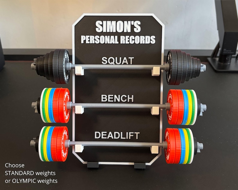 May include: A black and white sign displays "SIMON'S PERSONAL RECORDS" with three barbell sets for Squat, Bench, and Deadlift. The barbells have black or colourful weight plates. Text at the bottom reads "Choose STANDARD weights or OLYMPIC weights."