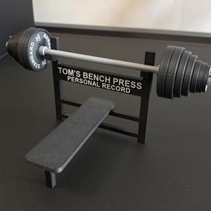 K&ouml;nnte beinhalten: Eine Miniatur-Hantelbank in Schwarz mit Langhantel und Gewichten. Die Langhantel hat auf jeder Seite Gewichte und der Text "STANDARD BAR" ist sichtbar. Auf der Bank steht der Text "TOM'S BENCH PRESS PERSONAL RECORD".