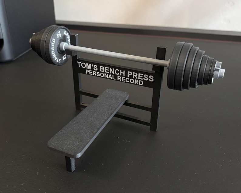 K&ouml;nnte beinhalten: Eine Miniatur-Hantelbank in Schwarz mit Langhantel und Gewichten. Die Langhantel hat auf jeder Seite Gewichte und der Text "STANDARD BAR" ist sichtbar. Auf der Bank steht der Text "TOM'S BENCH PRESS PERSONAL RECORD".