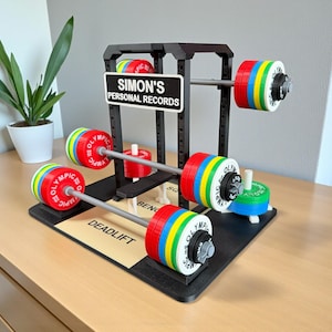 Puede incluir: Un juego de halterofilia en miniatura con un marco negro y pesas coloridas. El conjunto incluye un letrero que dice "SIMON'S PERSONAL RECORDS" y la palabra "DEADLIFT" en la base. Las pesas son rojas, amarillas, azules y verdes.