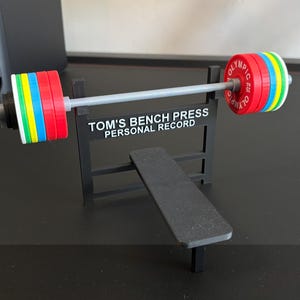K&ouml;nnte beinhalten: Ein Miniatur-Gewichtheberset mit Langhantel und Bank. Die Langhantel hat farbige Gewichtsscheiben in Rot, Blau, Gr&uuml;n und Gelb. Der Text auf dem St&auml;nder lautet "TOM'S BENCH PRESS PERSONAL RECORD".