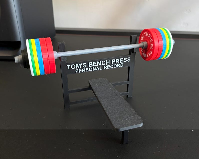 K&ouml;nnte beinhalten: Ein Miniatur-Gewichtheberset mit Langhantel und Bank. Die Langhantel hat farbige Gewichtsscheiben in Rot, Blau, Gr&uuml;n und Gelb. Der Text auf dem St&auml;nder lautet "TOM'S BENCH PRESS PERSONAL RECORD".