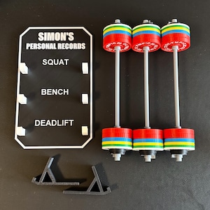 May include: Three miniature barbells with red, blue, yellow, and green weight plates. A black sign displays "SIMON'S PERSONAL RECORDS" and categories for squat, bench, and deadlift. Two black stands are also visible.