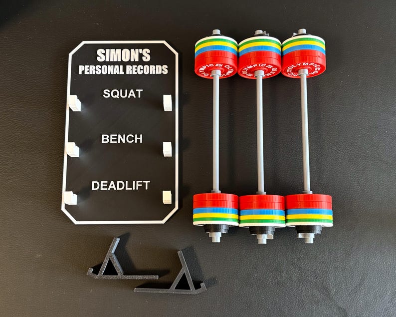 May include: Three miniature barbells with red, blue, yellow, and green weight plates. A black sign displays "SIMON'S PERSONAL RECORDS" and categories for squat, bench, and deadlift. Two black stands are also visible.