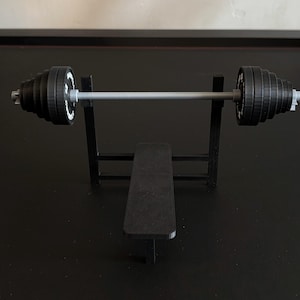 K&ouml;nnte beinhalten: Ein Miniatur-Hantelbank-Set mit einer schwarzen Bank und einem Gestell, das eine Langhantel mit schwarzen Gewichtsscheiben tr&auml;gt. Die Gewichte haben wei&szlig;e Markierungen. Die Langhantel ist silberfarben.