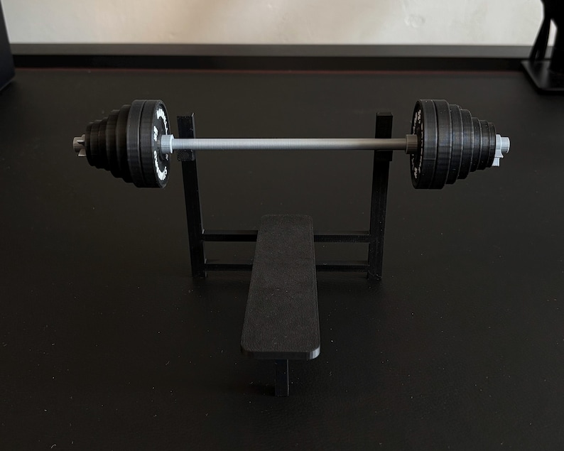 K&ouml;nnte beinhalten: Ein Miniatur-Hantelbank-Set mit einer schwarzen Bank und einem Gestell, das eine Langhantel mit schwarzen Gewichtsscheiben tr&auml;gt. Die Gewichte haben wei&szlig;e Markierungen. Die Langhantel ist silberfarben.