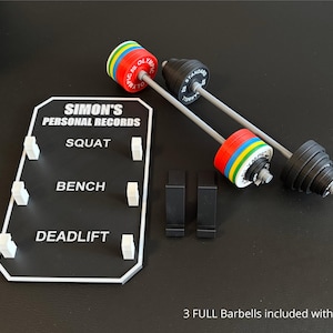 May include: Miniature barbell set with a record board. The barbells have black and silver weights with colourful plates. The record board is black with white text that reads "SIMON'S PERSONAL RECORDS" and lists "SQUAT", "BENCH", and "DEADLIFT".