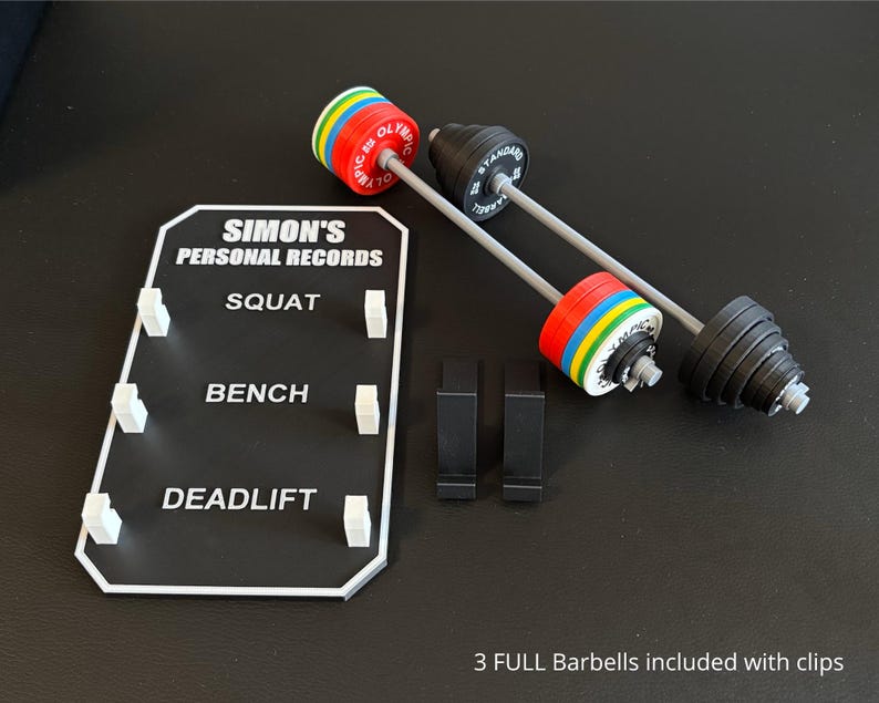 May include: Miniature barbell set with a record board. The barbells have black and silver weights with colourful plates. The record board is black with white text that reads "SIMON'S PERSONAL RECORDS" and lists "SQUAT", "BENCH", and "DEADLIFT".