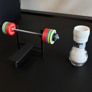 K&ouml;nnte beinhalten: Eine Miniatur-Langhantel mit bunten Gewichtsscheiben und einer kleinen Bank, neben einem durchsichtigen Salzstreuer mit wei&szlig;em Deckel. Die Langhantel hat rote, gelbe, blaue und gr&uuml;ne Scheiben. Der Salzstreuer ist mit wei&szlig;em Salz gef&uuml;llt.