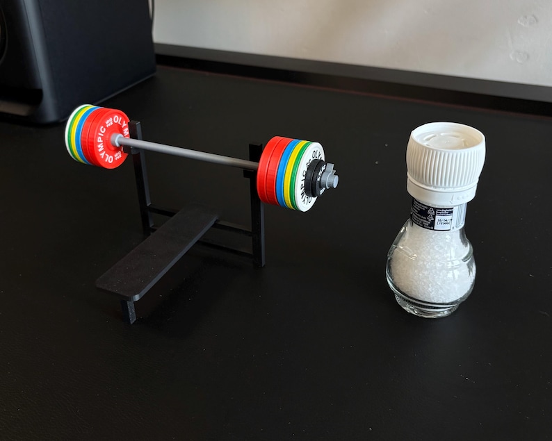 K&ouml;nnte beinhalten: Eine Miniatur-Langhantel mit bunten Gewichtsscheiben und einer kleinen Bank, neben einem durchsichtigen Salzstreuer mit wei&szlig;em Deckel. Die Langhantel hat rote, gelbe, blaue und gr&uuml;ne Scheiben. Der Salzstreuer ist mit wei&szlig;em Salz gef&uuml;llt.