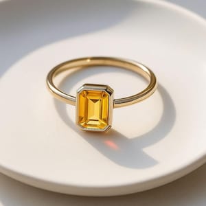Puede incluir: Un anillo de oro con una gema amarilla rectangular. El anillo tiene una banda delgada y un engaste biselado que enmarca la gema. El anillo se muestra sobre una superficie blanca, con iluminación suave y sombras.