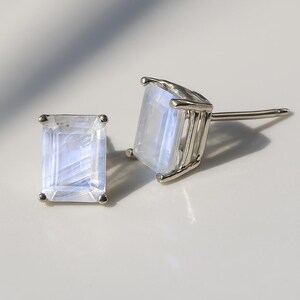 Puede incluir: Un par de pendientes de botón con piedras preciosas rectangulares e iridiscentes engastadas en metal plateado. Las piedras tienen un sutil color azul y blanco. Se muestran sobre una superficie clara.