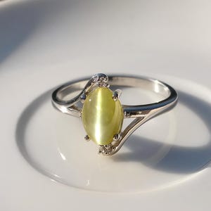 Puede incluir: Un anillo de plata con una piedra preciosa ovalada de color verde claro. El anillo tiene un diseño delicado y ondulado con pequeñas piedras de acento cerca de la gema principal. La piedra preciosa tiene un efecto ojo de gato.