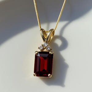 Puede incluir: Un collar con colgante de oro con una piedra preciosa rectangular de color rojo intenso. El colgante tiene tres pequeñas piedras preciosas transparentes y cuelga de una delicada cadena de oro. El engaste es de oro.