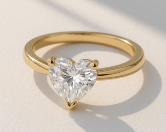 May include: A gold ring featuring a heart-shaped, clear gemstone. The band is a simple, polished gold. The gemstone is held in place by four prongs. The ring is set against a neutral background.