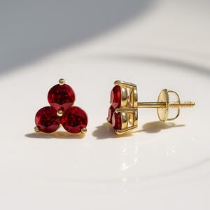 Puede incluir: Un par de pendientes de oro con tres gemas rojas redondas dispuestas en racimo. Los pendientes tienen un diseño clásico con cierre de rosca. Joyería elegante.