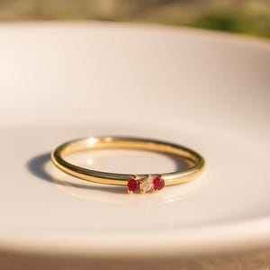 May include: A delicate gold ring featuring three gemstones: two red rubies flanking a central clear diamond. The ring's band is thin and polished, reflecting light. The gemstones are set close together, creating a subtle yet elegant design.
