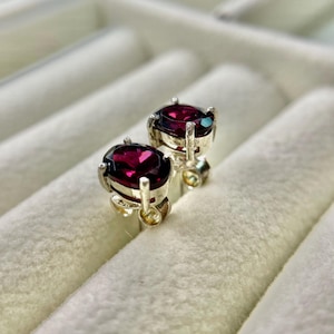 Puede incluir: Un par de pendientes de plata con piedras preciosas ovaladas de color rojo intenso. Los pendientes están engastados en metal plateado con cierre de poste y mariposa. El fondo es una superficie suave, blanca y texturizada.