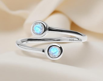 May include: A sterling silver bypass ring featuring two round, bezel-set moonstones with a blue iridescent glow. The band is stamped with 925 to indicate silver purity and includes a small maker's mark on the inner surface of the metal.