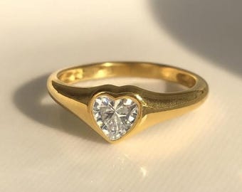 May include: A gold ring with a heart-shaped, clear gemstone. The ring has a smooth, polished band and a bezel setting that holds the gemstone securely. The ring is displayed on a white surface.