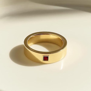 Può includere: Un anello dorato con una fascia piatta e una gemma rossa quadrata. L'anello ha una finitura lucida e un design minimalista. La gemma è incastonata nella fascia.